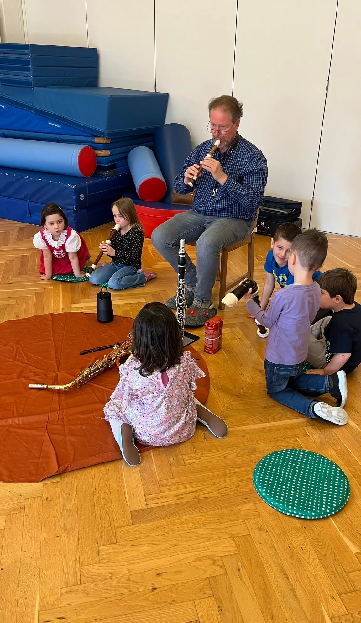 Ein Mann spielt eine Flöte mit Kindern um ihn herum. Einige Kinder halten Flöten. Eine Klarinette liegt auf dem Boden. Einige Kissen sind auf dem Boden. Eine Aufbewahrungsbox steht rechts.