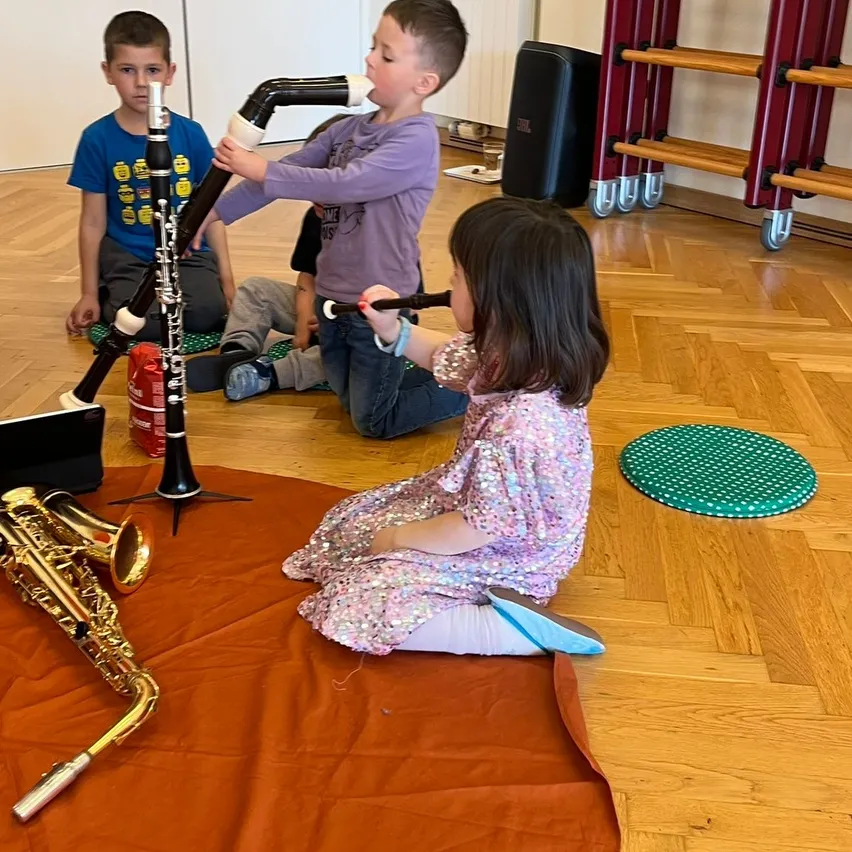 Drei Kinder spielen in einem Raum Musikinstrumente. Ein Junge bläst eine Klarinette, ein anderer Junge hält eine Flöte und ein Mädchen sitzt auf dem Boden mit einer Trompete vor sich.
