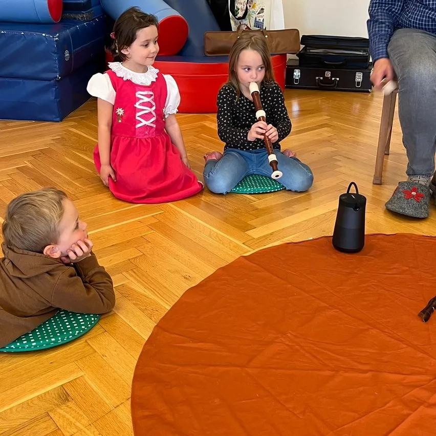 Zwei Mädchen und ein Junge sitzen auf dem Boden und sind in einer Musikstunde engagiert. Ein Mädchen spielt eine Blockflöte, während die anderen aufmerksam zuschauen. Ein Mann steht neben ihnen, und verschiedene Spielzeuge und eine Aktentasche sind im Hintergrund.