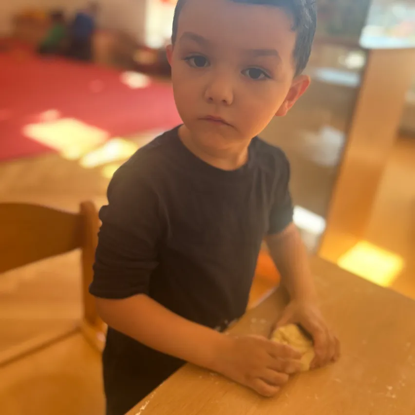Ein kleiner Junge in einem schwarzen T-Shirt sitzt an einem Holztisch und knetet konzentriert Teig.