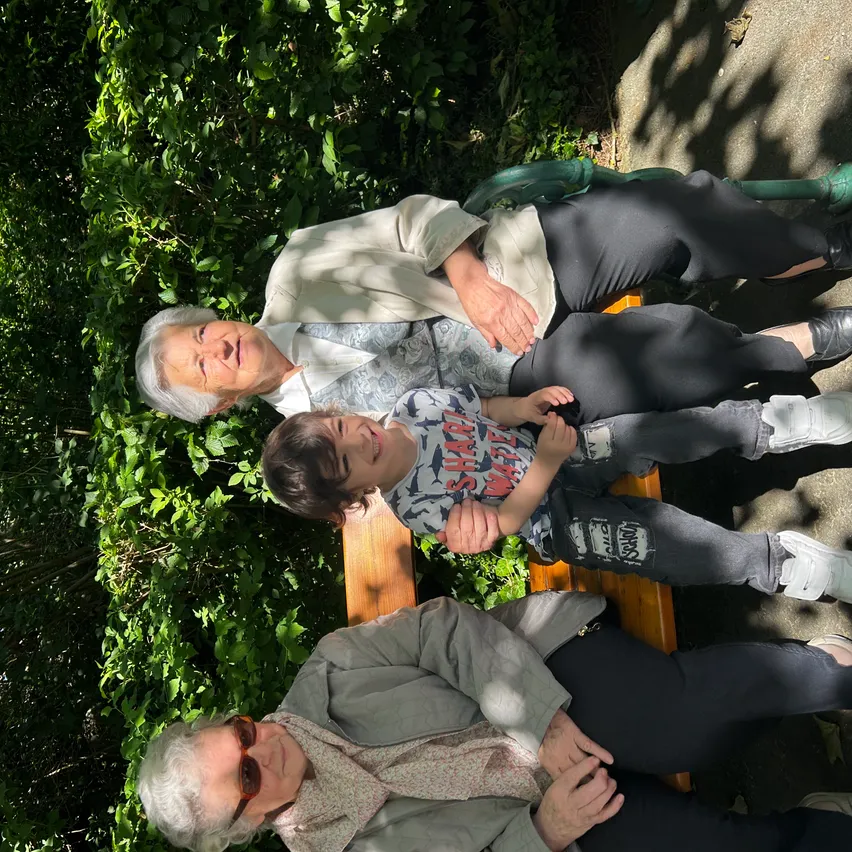 Drei ältere Frauen und ein junger Junge sitzen auf einer Bank in einem Garten mit grünen Pflanzen und Bäumen.
