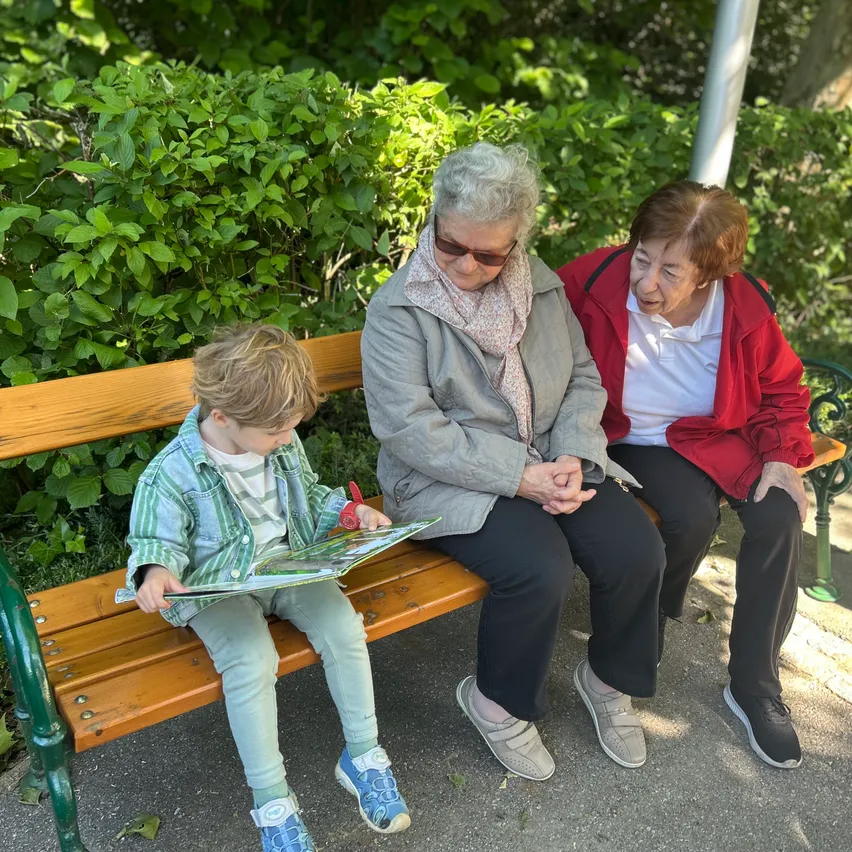 Drei Frauen und ein Junge sitzen auf einer Bank und lesen ein Buch. Der Junge liest laut vor, während die Frauen zuhören und lächeln.