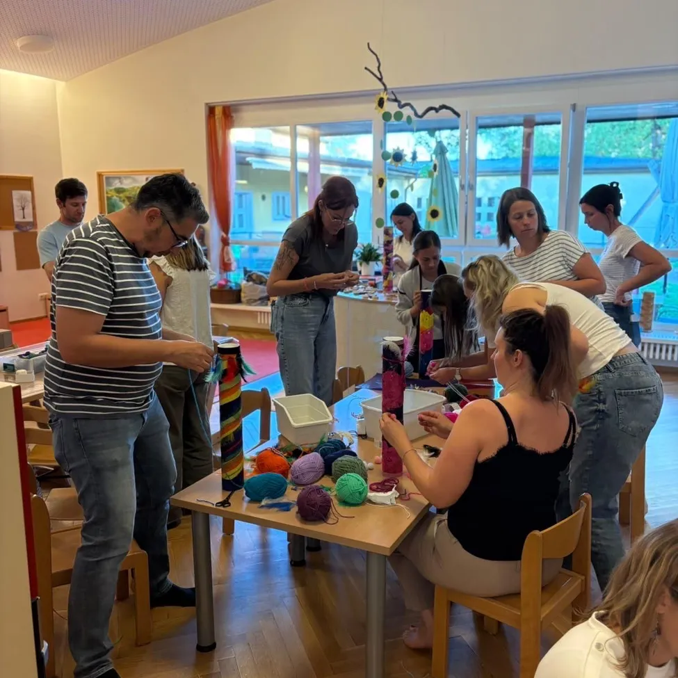 Eine Gruppe von Menschen versammelt sich in einem Raum und beschäftigt sich mit einer Bastelaktivität. Sie sitzen um einen Tisch herum mit verschiedenen Garnen und Stricknadeln. Der Raum hat große Fenster und einen dekorativen Sonnenblumenaufhänger.