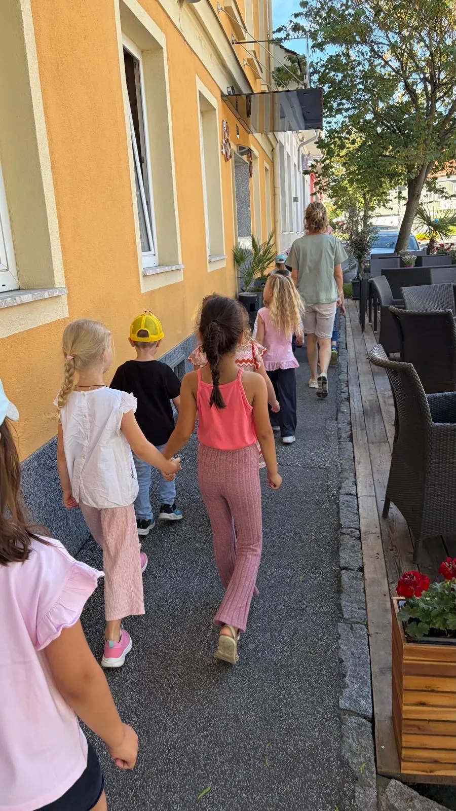 Eine Gruppe von Kindern geht Hand in Hand die Gehweg entlang. Ein Mädchen trägt ein rosa Oberteil und Hose. Ein Erwachsener folgt ihnen. Topfpflanzen und Stühle sind auf der Seite.