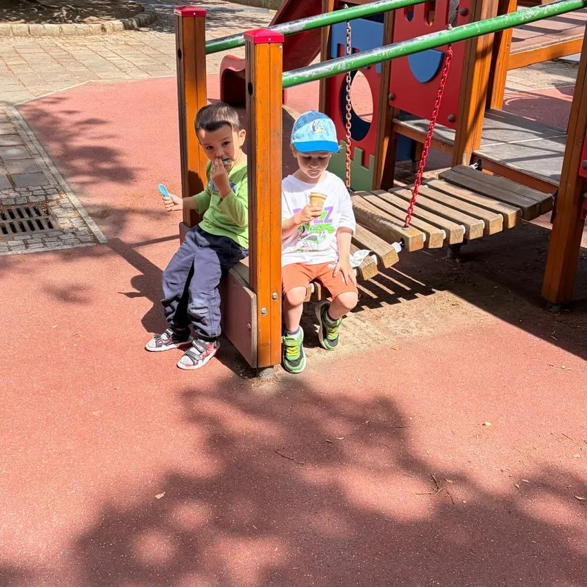 Zwei Jungen sitzen auf einer Bank im Spielplatz und essen Eiscreme und halten Eislutscher. Sie tragen beide Mützen und Turnschuhe.