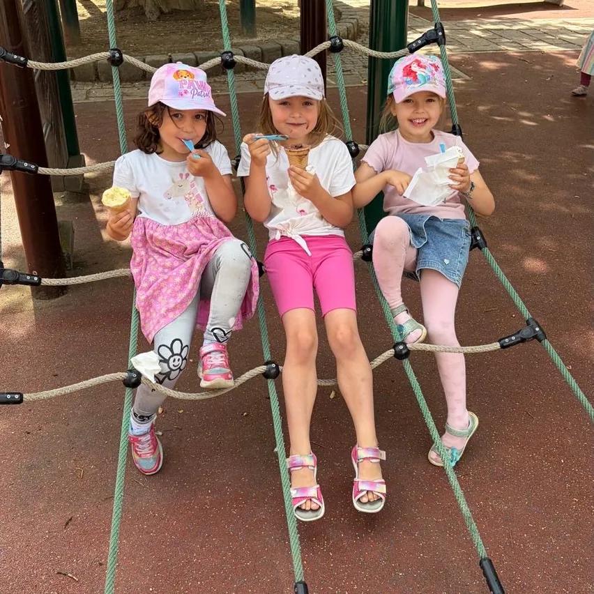 Drei junge Mädchen sitzen auf einem Kletternetz im Spielplatz. Zwei von ihnen essen Eis und eine hält ein Taschentuch. Sie tragen alle Mützen und Sandalen.