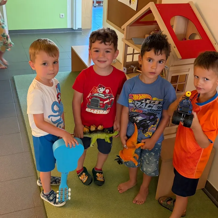 Vier junge Jungen stehen drinnen, halten Spielzeug und lächeln. Ein Dinosaurier-Spielzeug ist prominent zu sehen. Ein Holzhaus und ein Teppich sind im Hintergrund.