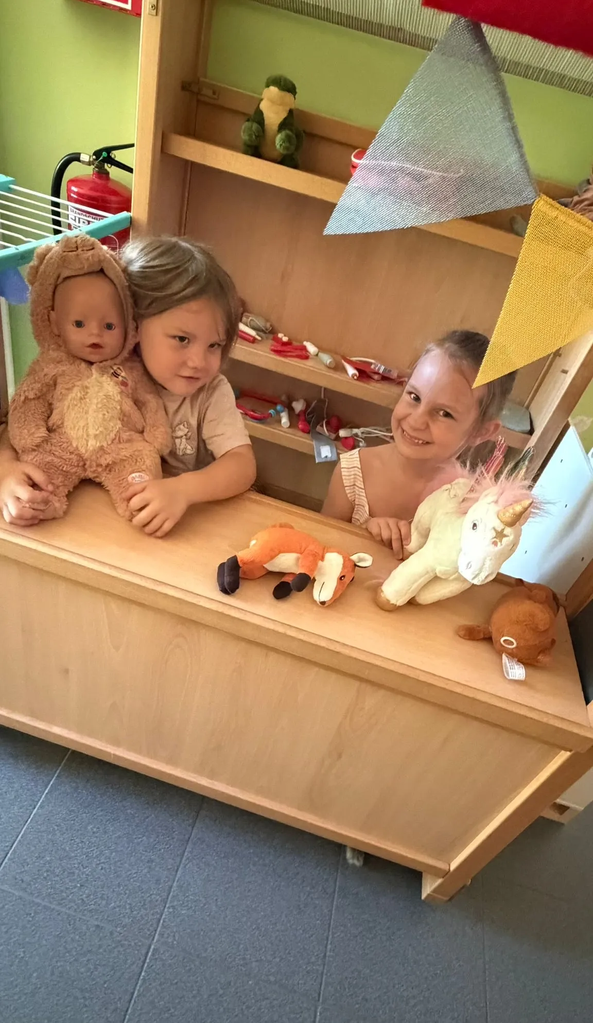 Zwei Kinder spielen in einem Raum mit Spielzeug. Ein Junge hält einen Teddybären. Ein Mädchen sitzt hinter einem Schreibtisch mit einem Einhorn-Spielzeug.