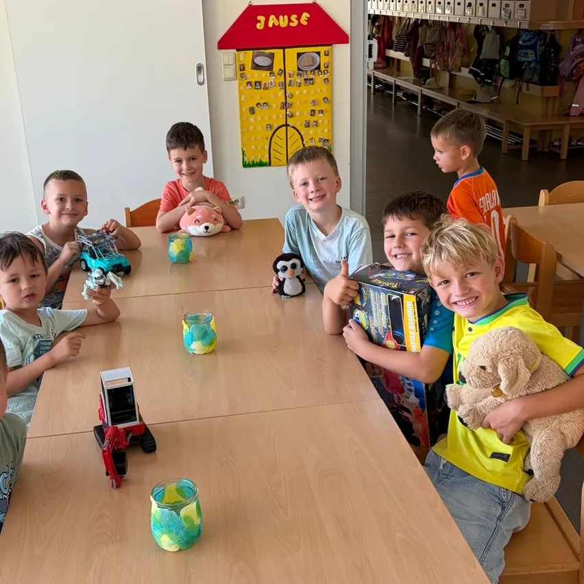 Eine Gruppe von Jungen in einem Raum mit Holztischen, die Spielzeug halten. Ein rotes Schild mit dem Wort 'JAUSE' ist an der Wand.