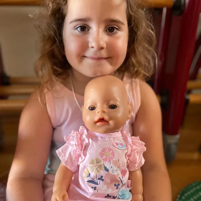 Ein junges Mädchen mit lockigem Haar hält eine rosa bekleidete Babypuppe mit Blumendesign. Das Mädchen lächelt und sitzt auf dem Boden, während sich eine rote Struktur hinter ihr befindet.