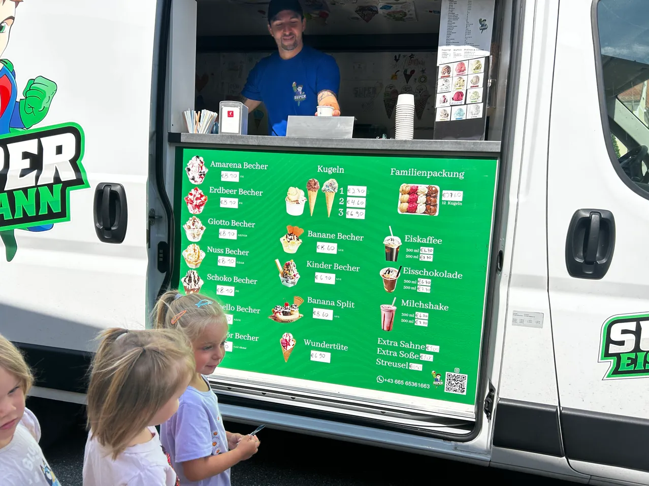 Ein Eiswagen mit Speisekarte, die verschiedene Geschmacksrichtungen wie Bananensplit, Milchshake usw. zeigt. Ein Mann ist drinnen. Drei junge Mädchen schauen sich die Speisekarte an.