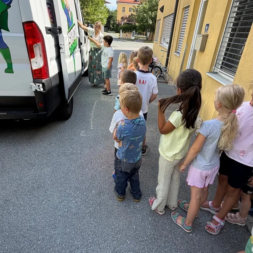 Mehrere Kinder und eine Erwachsene stehen um einen weißen Van mit einer grünen Superheldenfigur herum und schauen auf den Heck des Vans. Eine Frau steht neben dem Van.