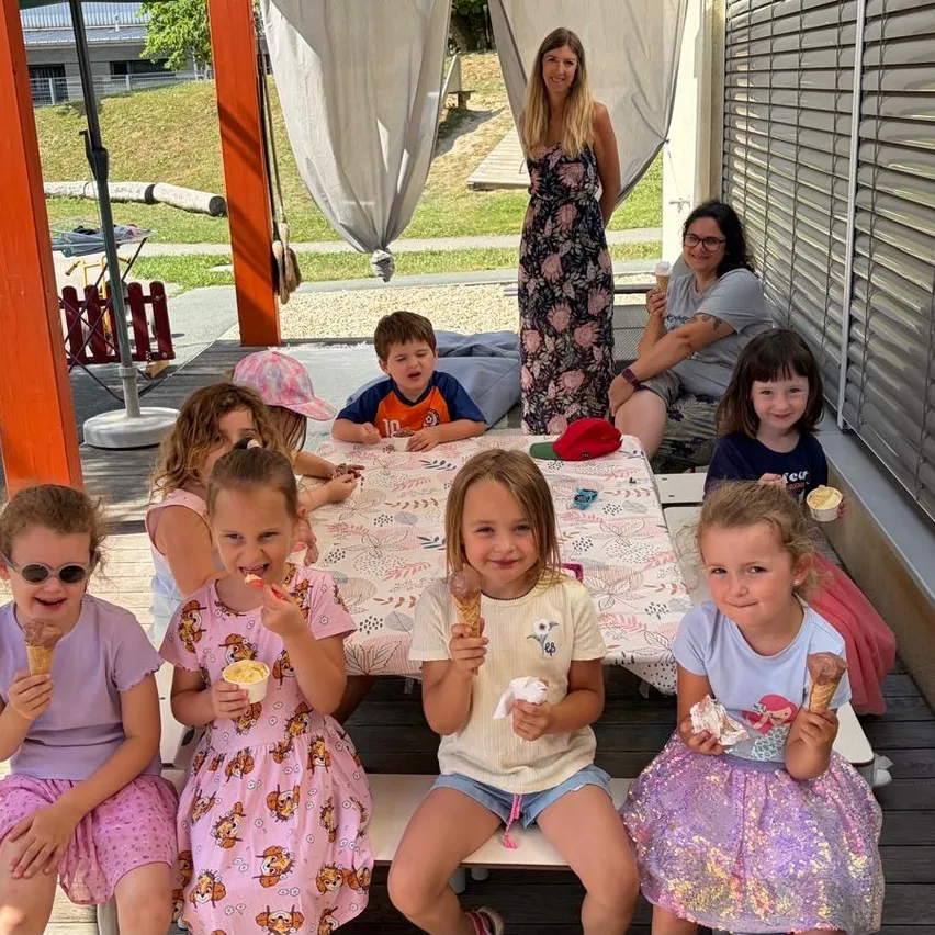 Eine Gruppe von Kindern und zwei Erwachsenen sitzen um einen Tisch und essen Eis auf einer Veranda. Eine Frau steht hinter ihnen.