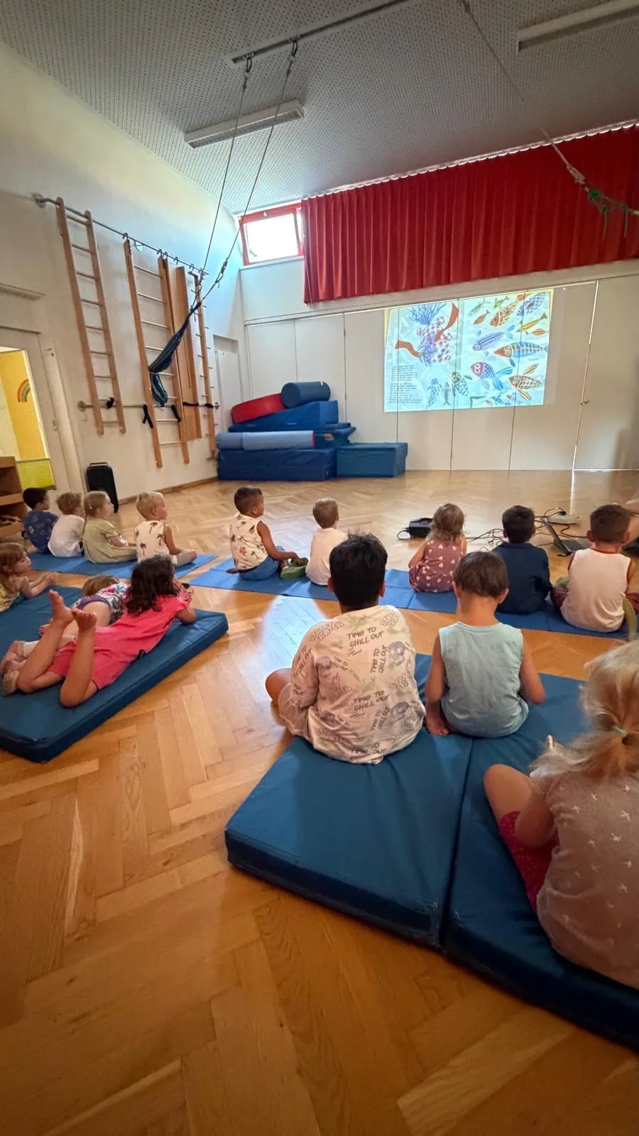 Eine Gruppe junger Kinder sitzt auf blauen Matten in einem Raum und schaut sich eine fischthematische Präsentation auf einer Projektionsleinwand an.