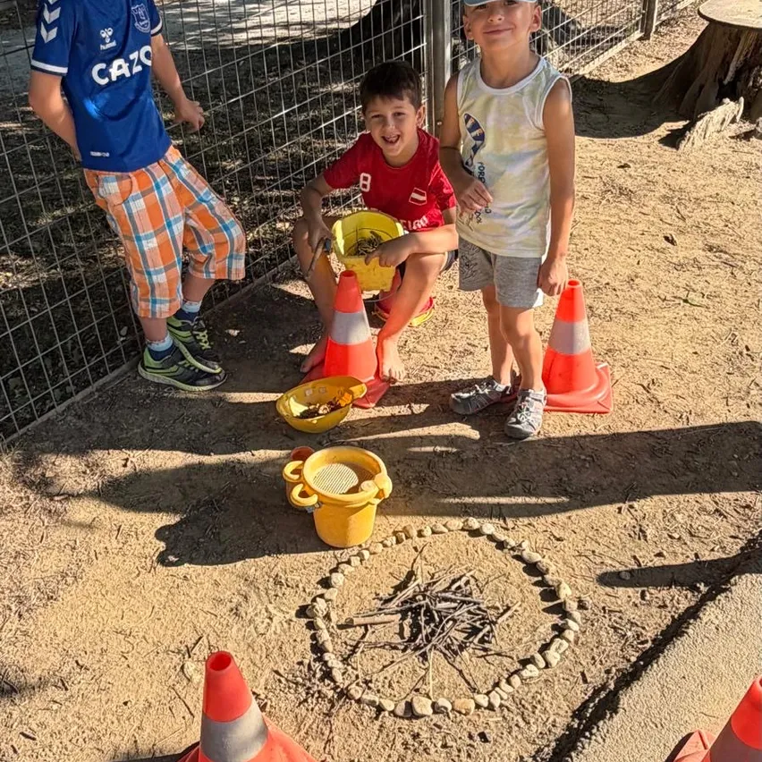Drei Jungen in einem Zoogehege, einer sitzt auf dem Boden mit einem gelben Eimer, ein anderer steht mit einem Eimer und der dritte steht neben einem Verkehrskegel.