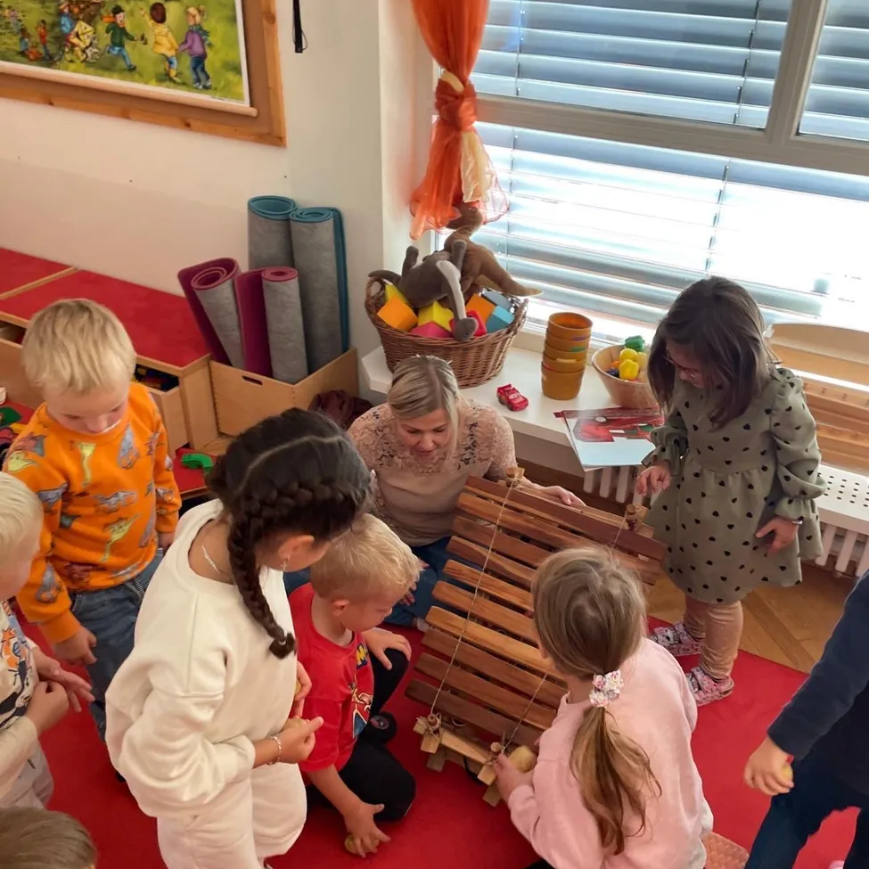 Eine Gruppe von Kindern und einer erwachsenen Frau spielen mit einem hölzernen Xylophon in einem Raum mit einem roten Teppich und einem Fenster mit Jalousien.