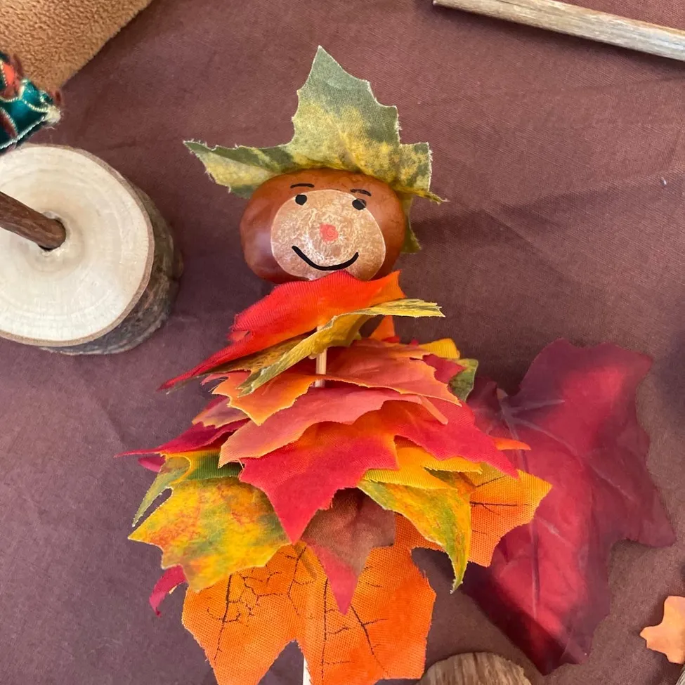 Eine handgefertigte Herbstdekoration mit einem lächelnden Gesicht auf einer braunen Nuss und einer Krone aus bunten Blättern.