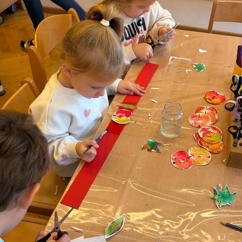 Drei Kinder sitzen an einem Tisch und sind in einer Kunstaktivität verwickelt. Ein Kind schneidet ein rotes Band, während die anderen beiden auf Papier malen. Der Tisch ist mit Plastik bedeckt und es gibt verschiedene Kunstmaterialien um sie herum.