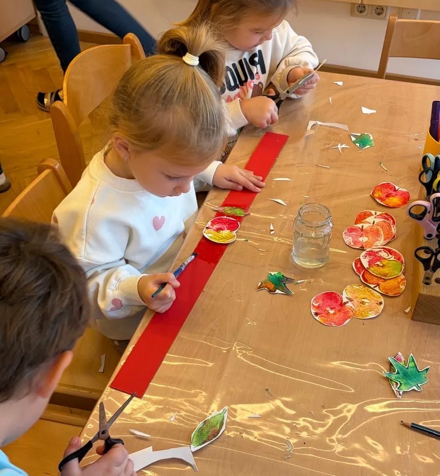 Drei Kinder sitzen an einem Tisch und sind in einer Kunstaktivität verwickelt. Ein Kind schneidet ein rotes Band, während die anderen beiden auf Papier malen. Der Tisch ist mit Plastik bedeckt und es gibt verschiedene Kunstmaterialien um sie herum.