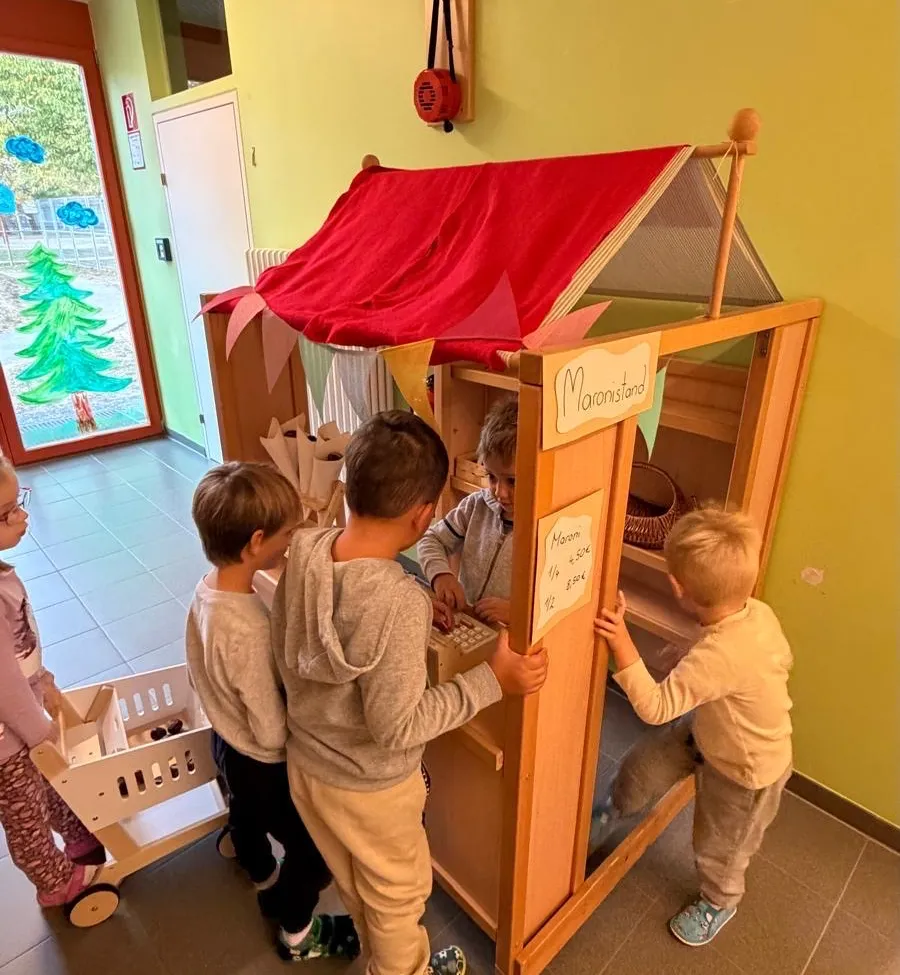 Eine Gruppe von Kindern spielt in einem hölzernen Spielhaus mit einem roten Dach und einem Schild, das 'Maronistand' steht. Sie schauen in das Spielhaus hinein.