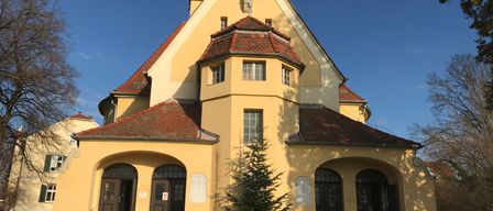 A yellow church building with red roof, a statue of a person on the gable, and three arched doors at the front.