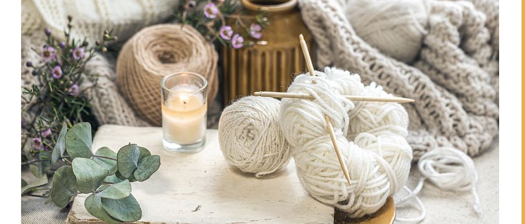 Ein gemütliches Plakat für ein Handarbeits-Café mit einer Platte aus Wolle, Stricknadeln, einer Kerze und einer gestrickten Decke. Der Text lautet 'Handarbeits-Cafe'.