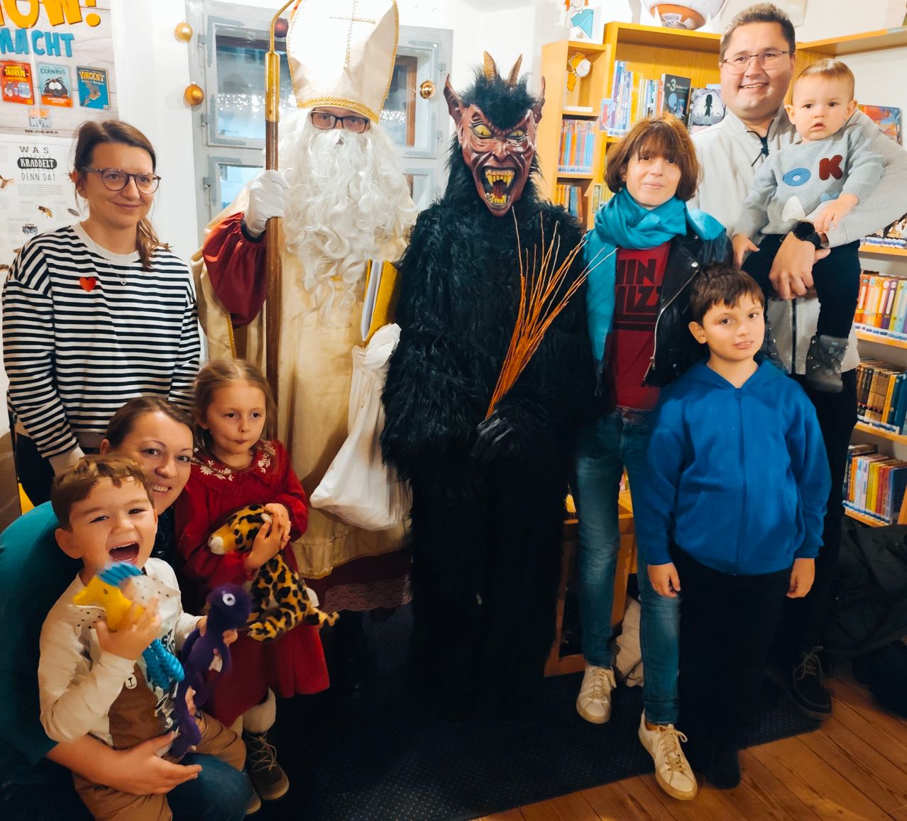 Ein Familienfoto in einer Bibliothek mit zwei Erwachsenen in Kostümen, einer als Weihnachtsmann und der andere als Teufel, stehend mit Kindern, Spielzeug und einem Baby auf dem Schoß eines Mannes.