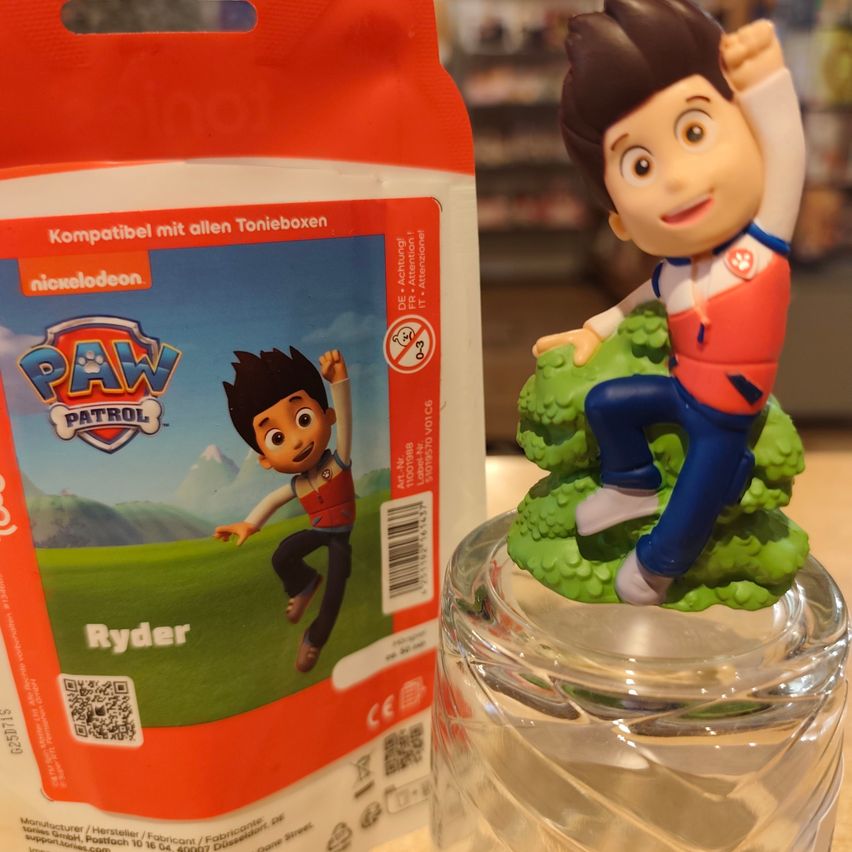 A Ryder Paw Patrol figurine is displayed on a glass surface next to its packaging. The packaging is orange with white text. The figurine is on a green bush.