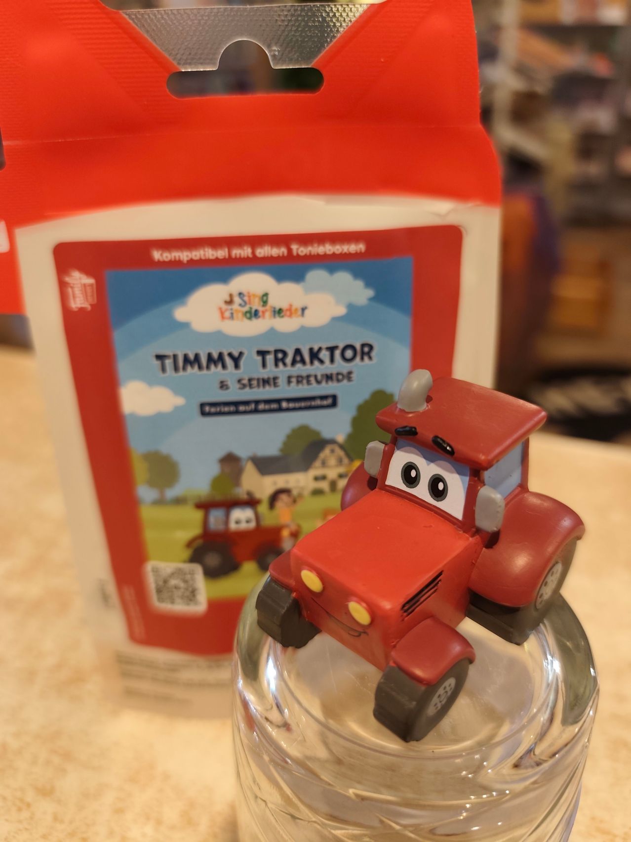 A small red toy tractor sits atop a glass cup, with a red and white bag of children's storybook behind it. The bag features Timmy Traktor and his friends in a countryside setting.