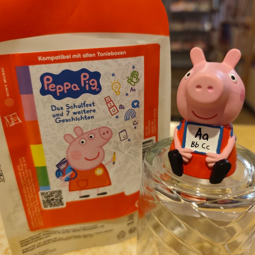A Peppa Pig figurine holding a sign with alphabet letters is next to a Peppa Pig storybook. The book is titled Das Schulfest und 7 weitere Geschichten.