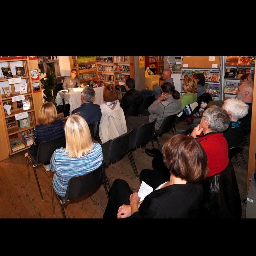 Ein Publikum hört einem sitzenden Mann in einer Buchhandlung zu, mit Bücherregalen und einem Tisch im Hintergrund.