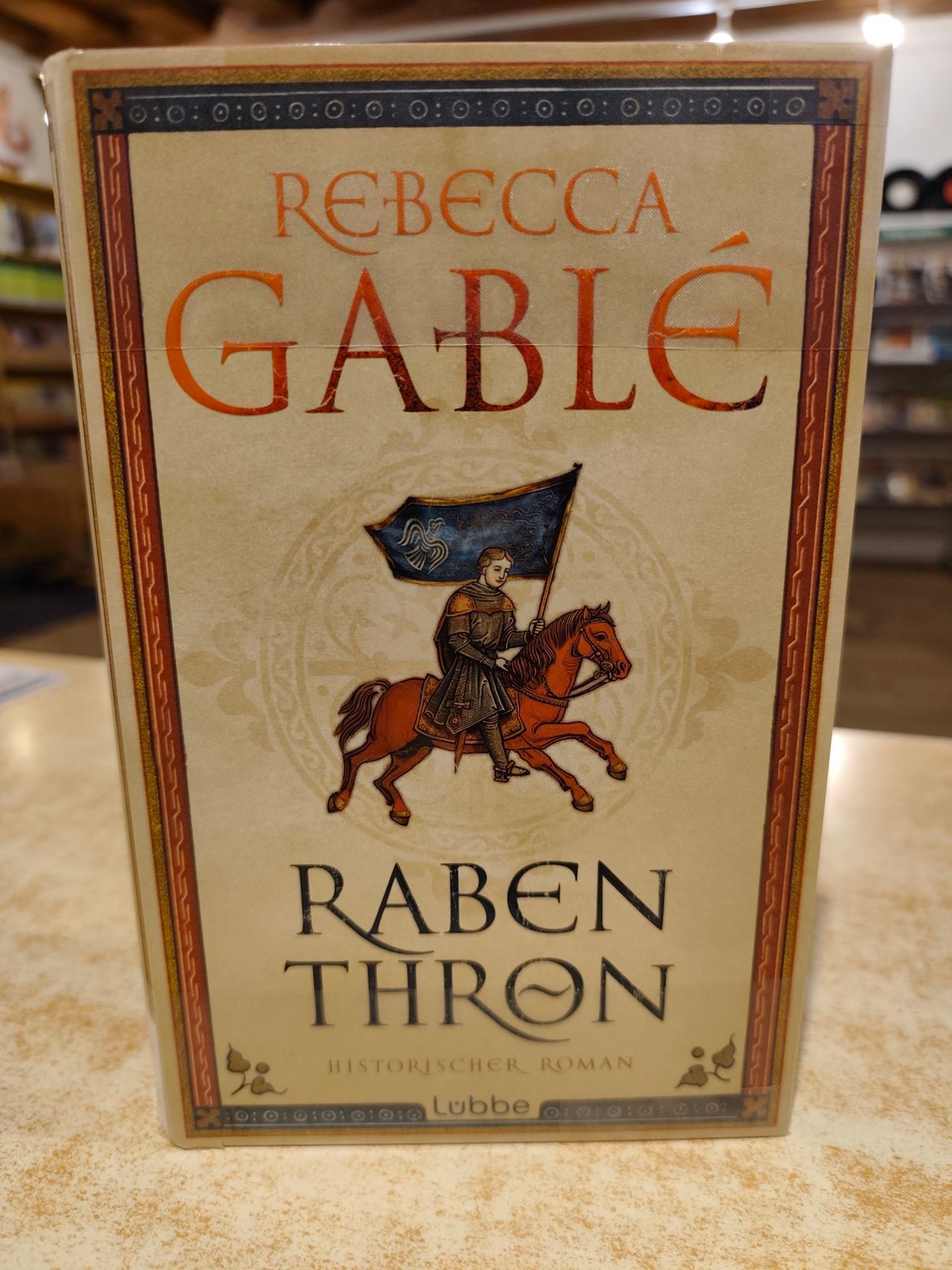 Ein Buchcover zeigt einen Ritter auf einem Pferd mit einer Fahne. Der Titel lautet 'Raben Thron' in großen Buchstaben. Die Autorin ist Rebecca Gable.