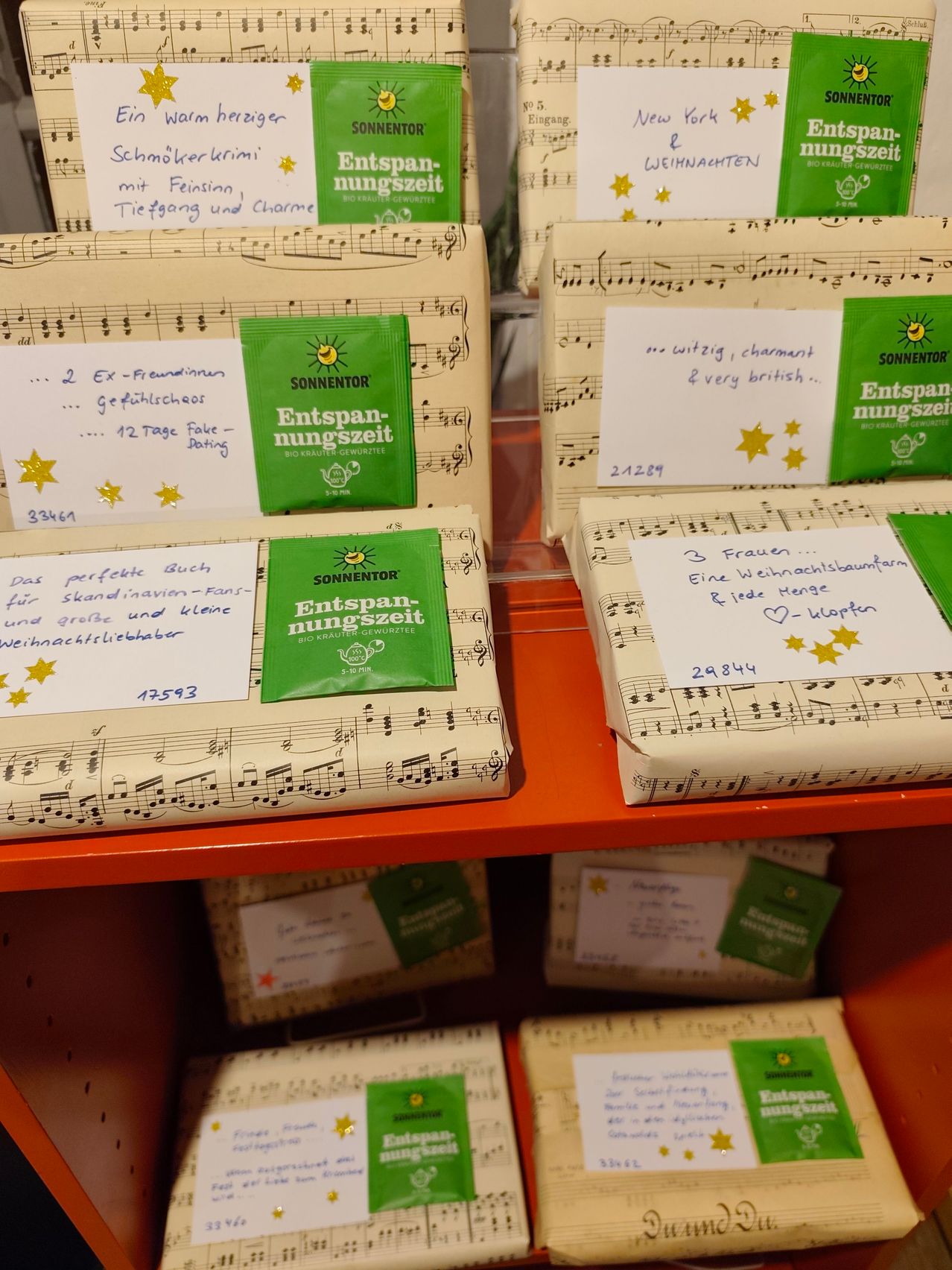 Ein Regal zeigt mehrere verpackte Geschenke mit Musiknoten und handgeschriebenen Notizen. Jedes Paket hat ein grünes Etikett mit dem Text 'Entspannungszeit' und einem Sonnensymbol. Die Geschenke sind in einer Reihe angeordnet, und einige sind teilweise ausgepackt.