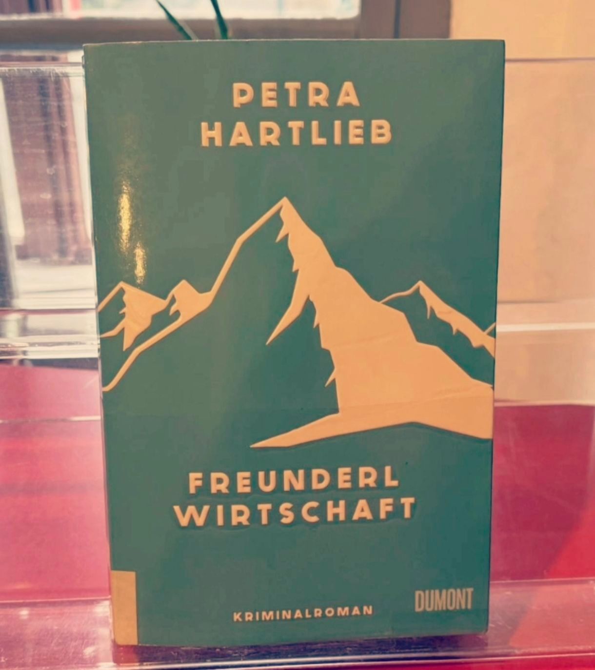 Ein grünes Buch mit dem Titel 'Freunderl Wirtschaft' von Petra Hartlieb wird auf einem Glasregal mit rotem Hintergrund ausgestellt. Das Buchcover zeigt eine goldene Illustration eines Berges.