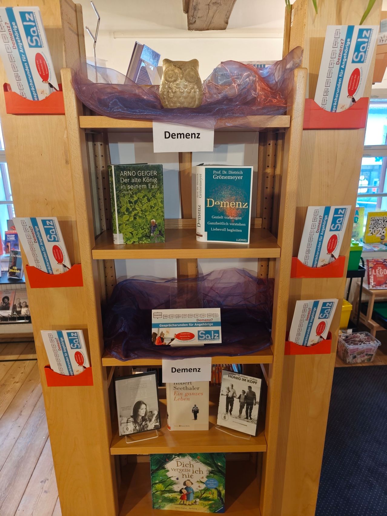 Ein hölzerne Bücherregal mit Büchern über Demenz, eines mit dem Titel 'Demenz' von Prof. Dr. Dietrich Grönemeyer und ein anderes von Arno Geiger mit dem Titel 'Der alte König in seinem Exil.' Es gibt eine Plastikabdeckung auf dem obersten Regal und ein Etikett mit der Aufschrift 'Demenz'.