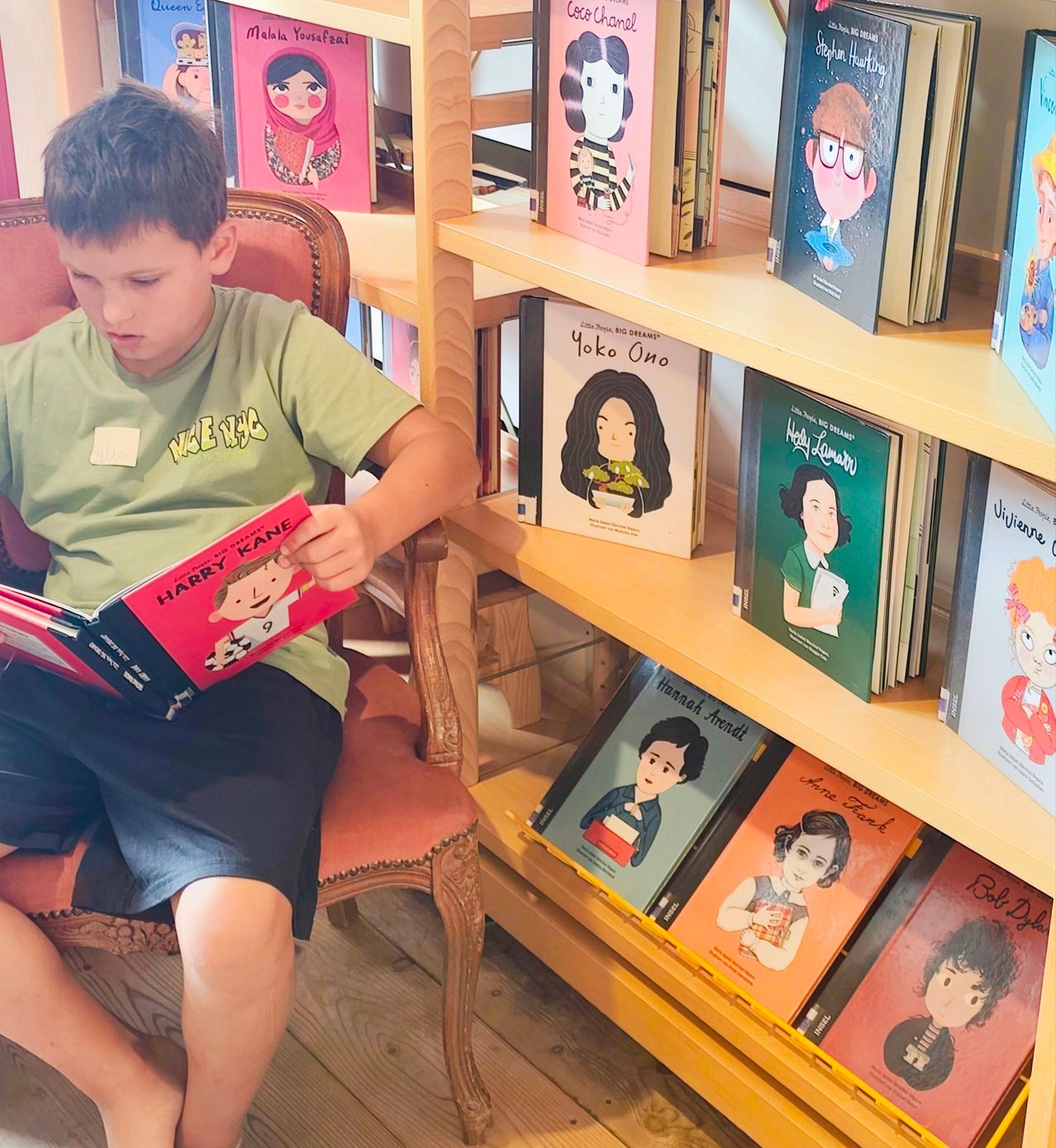 A young boy is sitting on a chair and reading a book titled 'Harry Kane' in a room with bookshelves filled with colorful books.