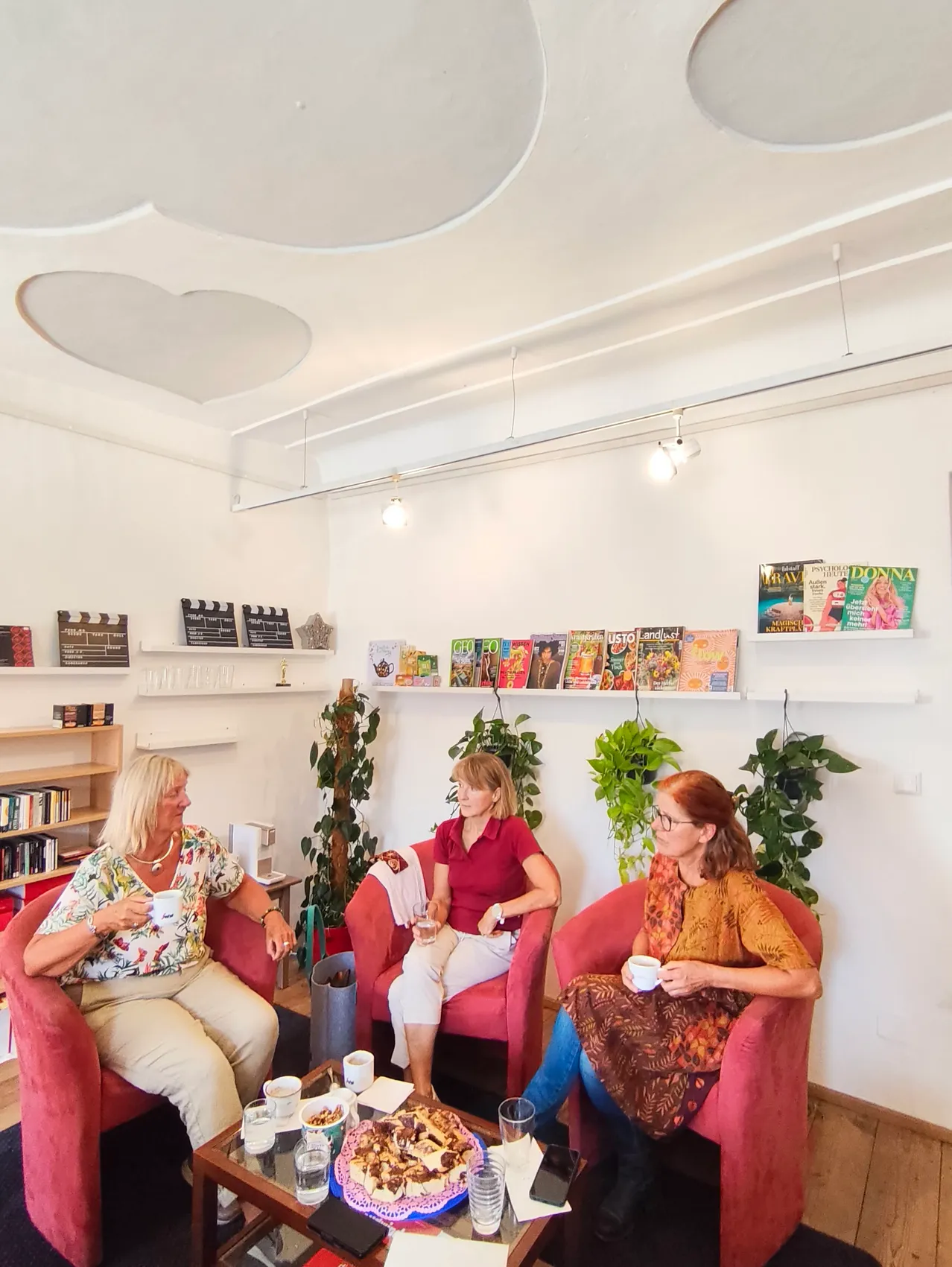Vier Frauen sitzen in roten Stühlen in einem Raum, halten Tassen und unterhalten sich. Regale mit Büchern und Zeitschriften sind an der Wand, und ein herzförmiges Design ist an der Decke.