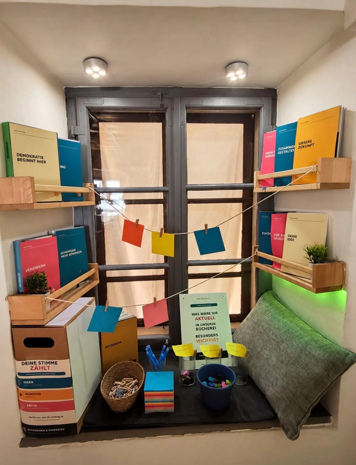 Eine gemütliche Leseecke mit Holzregalen, die bunte Bücher halten, einem Fenster mit Vorhang und einer kleinen Topfpflanze. Ein Schild steht: 'Was war die aktuelle in unserer Bucherei besonders wichtig?' mit bunten Haftnotizen, die an einer Schnur hängen.