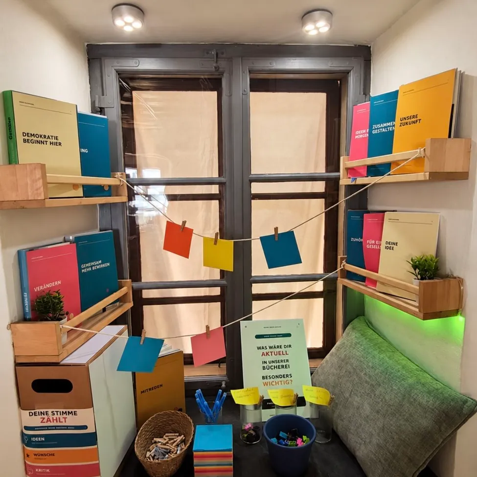 A cozy reading nook with wooden shelves holding colorful books, a window with a curtain, and a small potted plant. A sign reads 'Was war die aktuelle in unserer Bucherei besonders wichtig?' with colorful sticky notes hanging from a string.