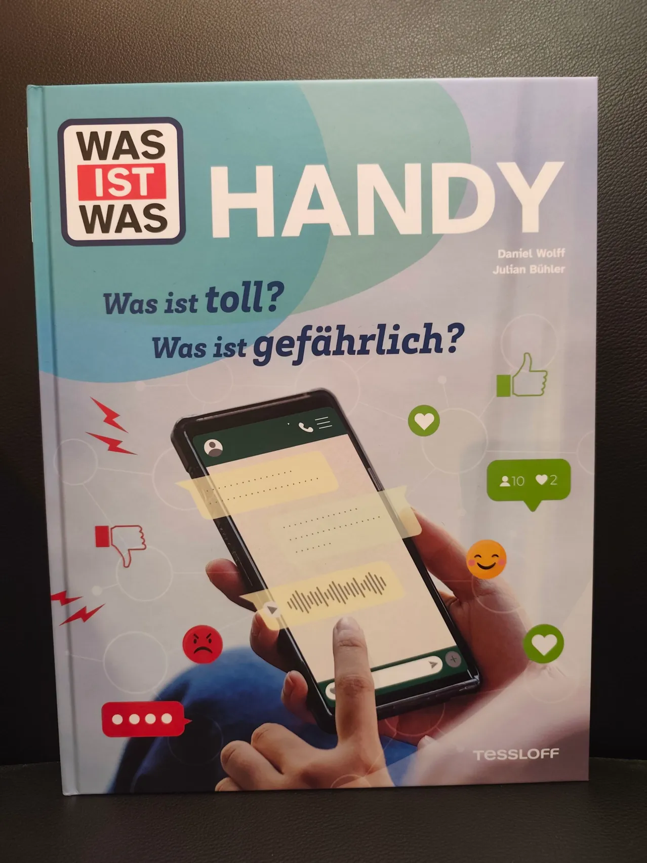 Ein Buchcover mit dem Titel 'Was ist Handy', das eine Hand mit einem Smartphone zeigt. Das Cover zeigt ein Smartphone mit Daumen hoch, Daumen runter, Herz und ärgerliches Gesichtssymbol.