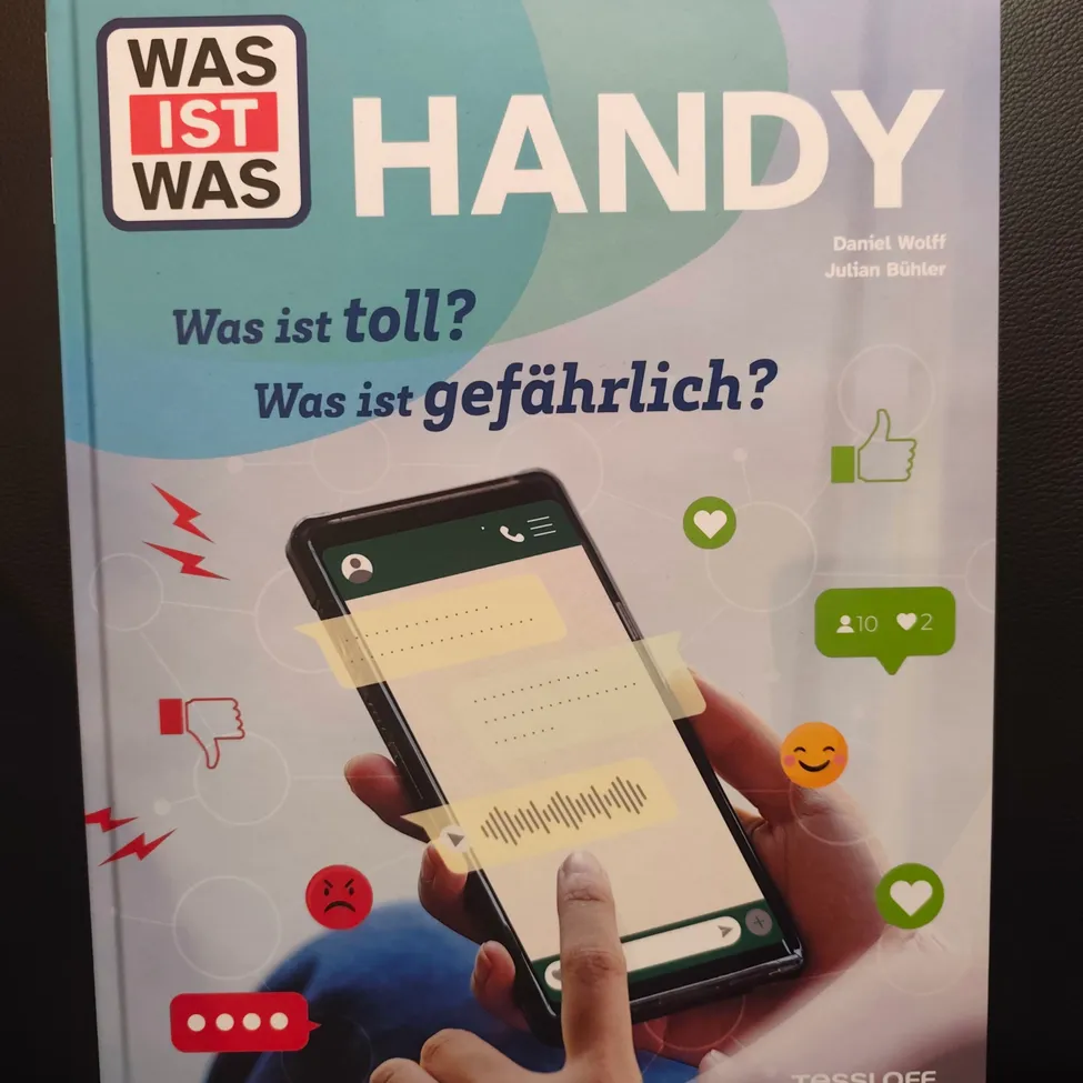 A book cover titled 'Was ist Handy' with a hand holding a smartphone. The cover features a smartphone with a thumbs-up, thumbs-down, heart, and angry face icons.
