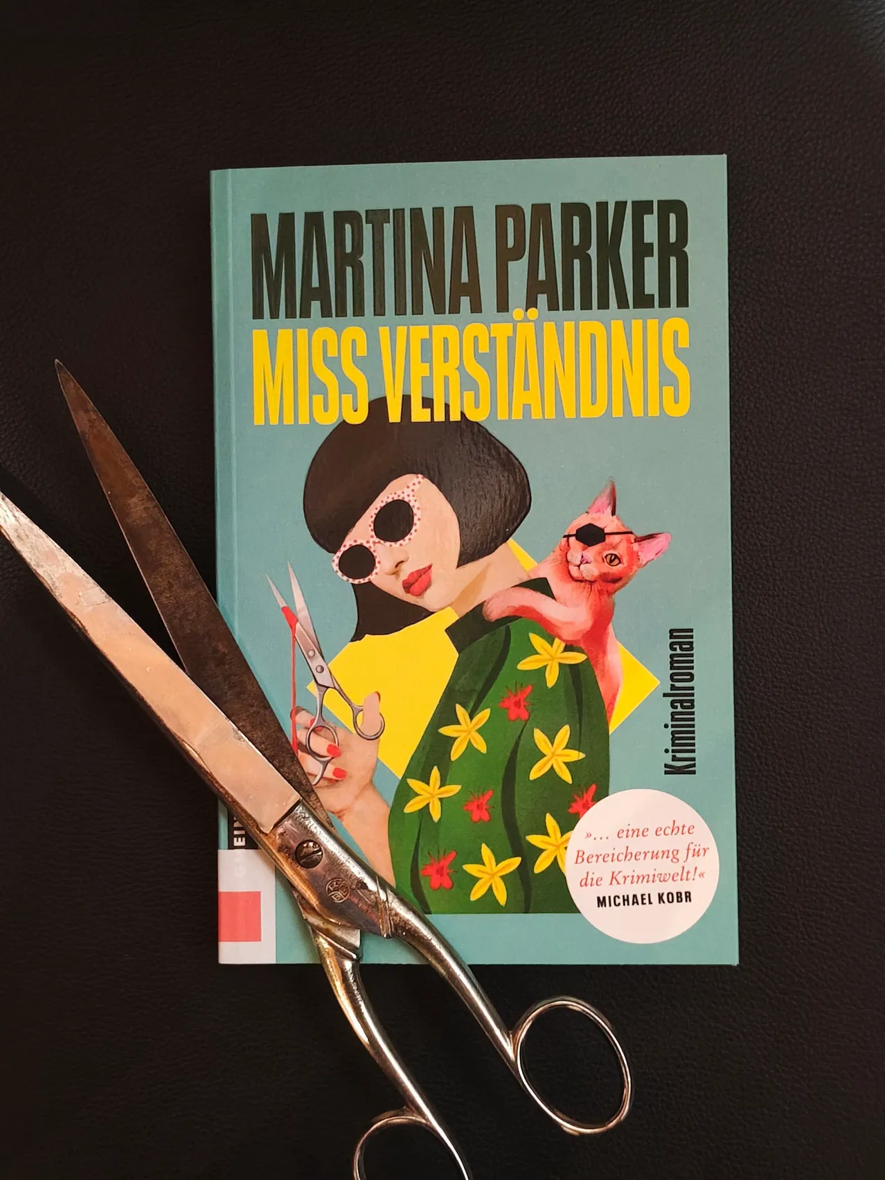 Ein Buch mit dem Titel 'Martina Parker Miss Verstandnis' mit einer Frau in einem gelben Kleid und einer Katze auf ihrer Schulter. Ein Paar Scheren schneidet das Buchcover.