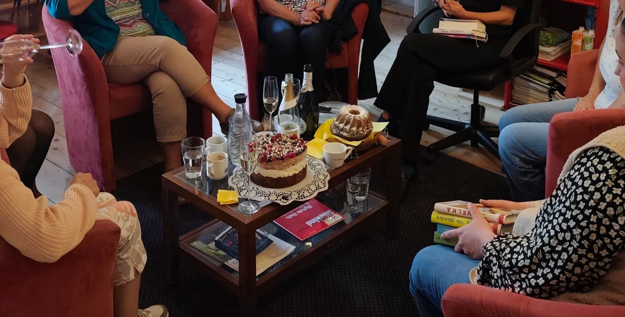 Eine Gruppe von Frauen sitzt in einem Raum, einige unterhalten sich und andere lesen Bücher. In der Mitte steht ein Tisch mit einem Kuchen, Weingläsern und Büchern.