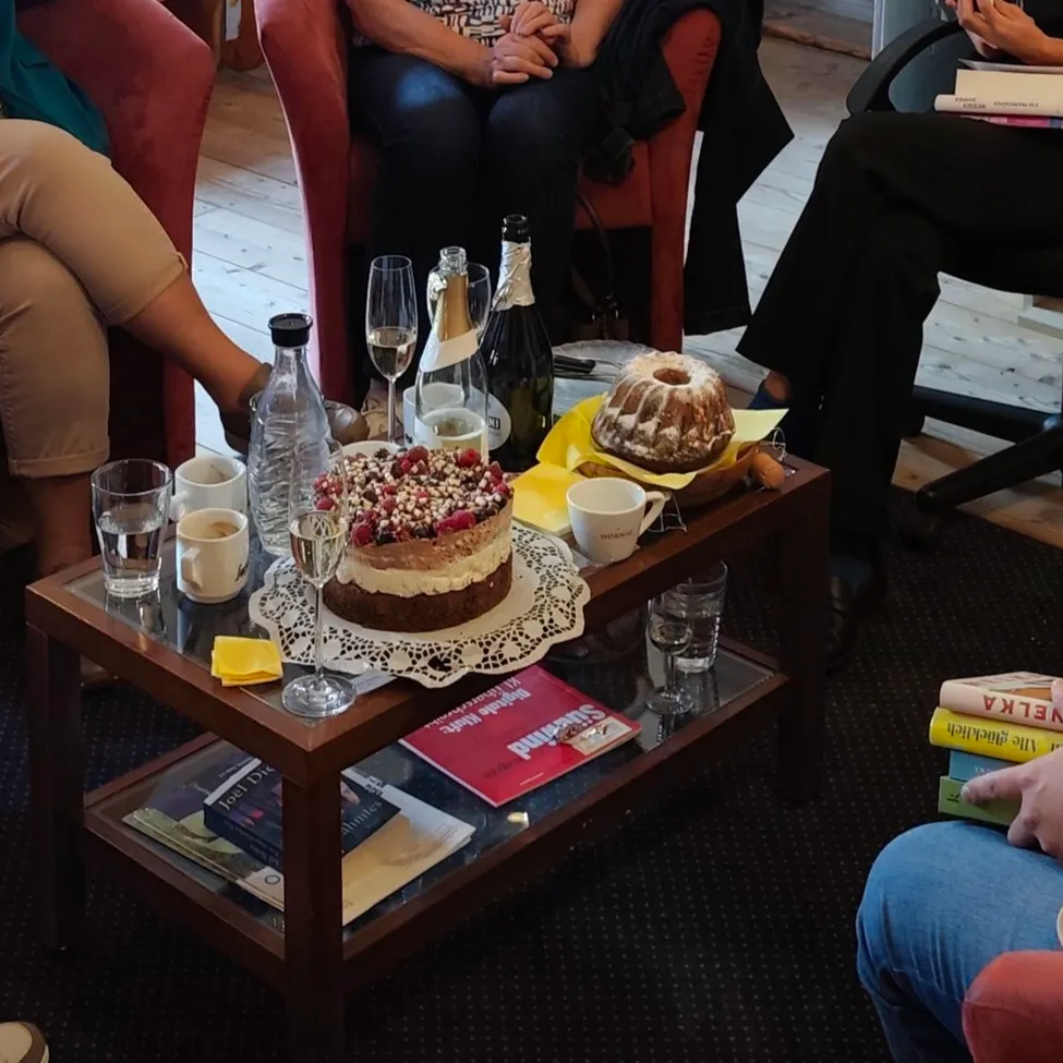 A group of women are sitting in a room, some are talking and others are reading books. There is a table with a cake, wine glasses, and books in the center.