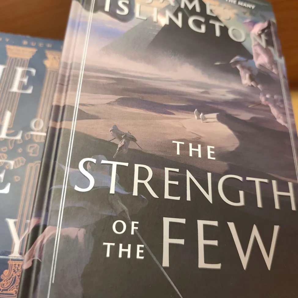 The book cover of 'The Strength of the Few' by James Islington. The cover features a desert landscape with a pyramid in the background, two figures in the foreground, and a sword in the middle.