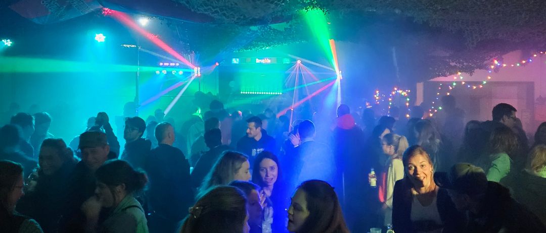 Ein überfüllter Nachtclub mit grünen und roten Laserlichtern, einer lächelnden Frau am Tisch und einer festlichen Atmosphäre.