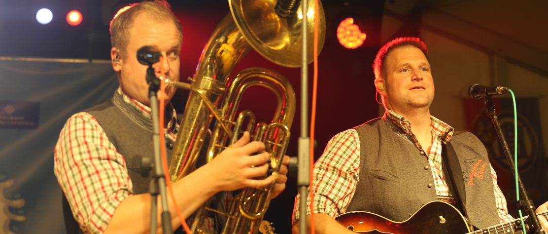 Zwei Männer auf der Bühne, einer spielt Tuba und der andere Gitarre. Beide stehen vor Mikrofonen. Die Bühne hat Scheinwerfer und Banner.
