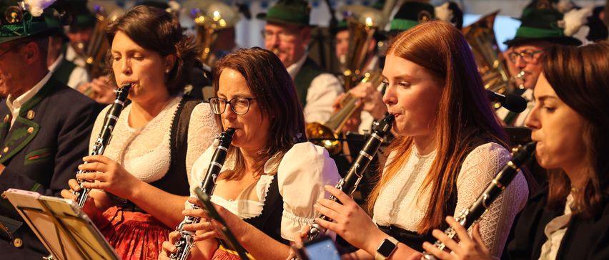 Eine Gruppe von Frauen in traditioneller Kleidung spielt Klarinetten bei einer Outdoor-Veranstaltung, mit Musikinstrumenten und Notenblättern im Hintergrund.