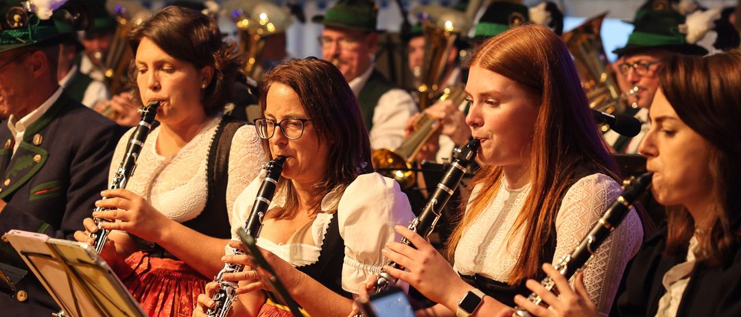 Eine Gruppe von Frauen in traditioneller Kleidung spielt Klarinetten bei einer Outdoor-Veranstaltung, mit Musikinstrumenten und Notenblättern im Hintergrund.