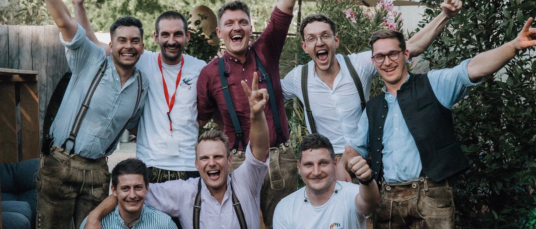 Eine Gruppe von Männern in traditioneller bayerischer Kleidung posiert für ein Foto, lächelt und macht Handzeichen in einer Außenumgebung mit Pflanzen und Sonnenschirmen im Hintergrund.